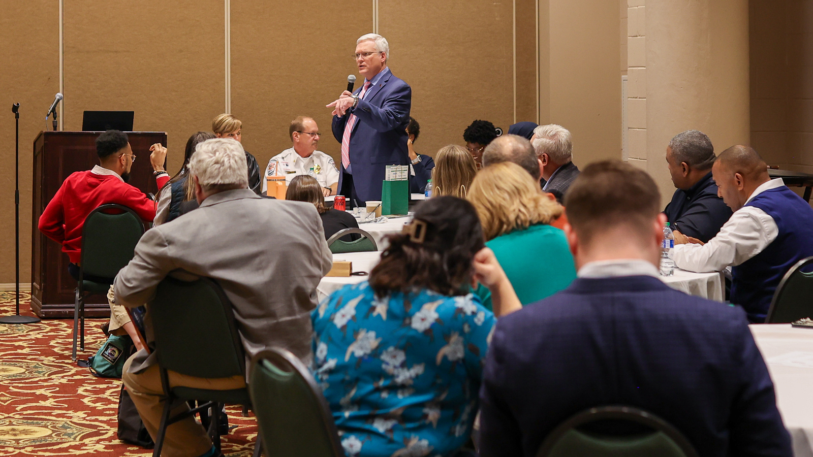 Man talking to a group of people in a class at a conference. He is holding a microphone. The audience is seated at tables.