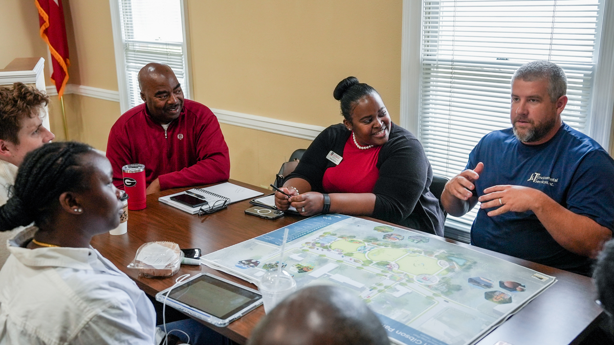 This image shows a group of people sitting around a table, engaged in a discussion. The table has a map on it, and there are other items on the table, such as phones, a tablet, and a red tumbler.